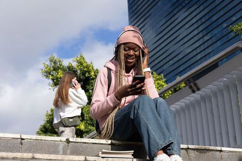 Smiling teen girl in pink hoodie listening to music and using smartphone on city steps