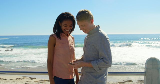 Man and woman stand by ocean, smiling while looking at phone. Potential usages include advertisements for travel destinations, relationship counseling, or lifestyle blogs promoting outdoor and beach activities.