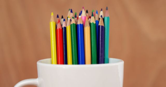 Close-up view of colorful pencils vertically arranged in a white mug. This image is ideal for use in educational contexts, promoting creativity and art-related content, classroom decoration ideas, or back-to-school themes. The vibrant colors of the pencils combined with the simplicity of the white mug and the neutral brown background create a visually appealing contrast.