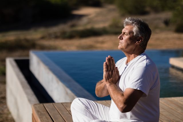 Senior man practicing yoga outdoors in sunlight, sitting cross-legged with hands in prayer position. Ideal for use in wellness, fitness, and mental health content, promoting relaxation, mindfulness, and healthy lifestyle.