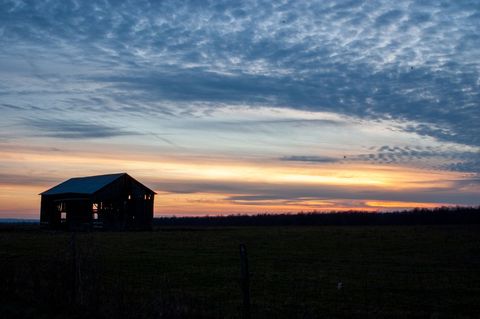 Weathered barn standing silhouetted against dramatic sunset sky over quiet farmland