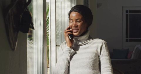 Woman talking on smartphone by sunlit window wearing cozy turtleneck sweater, smiling