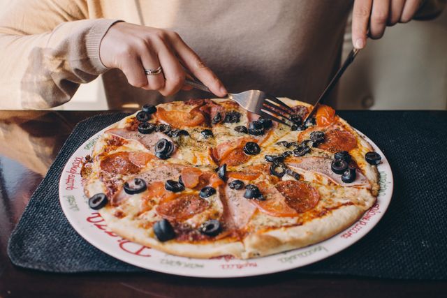 Shows a person dining at a restaurant while enjoying a delicious pepperoni and olive pizza. Perfect for use in advertisements for restaurants, food blogs, menu design, or culinary articles.