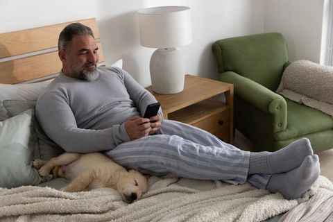 Mature man relaxing in bed with sleeping puppy while scrolling smartphone