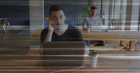 Young man talking on phone while working on laptop at coworking table with coffee cup