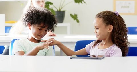 Children sharing tablet and touching hands in bright classroom, friendship and teamwork