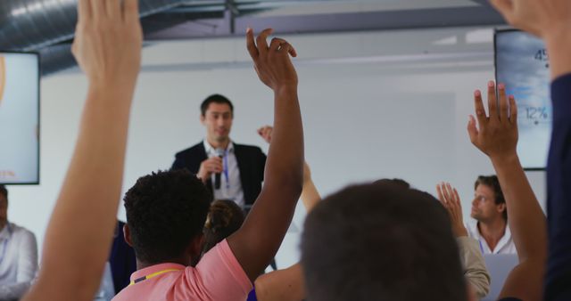 Front view of a young Caucasian businessman standing and talking to the audience at a business conference, seen through the raised arms of audience members who want to ask questions. Rack focus shot