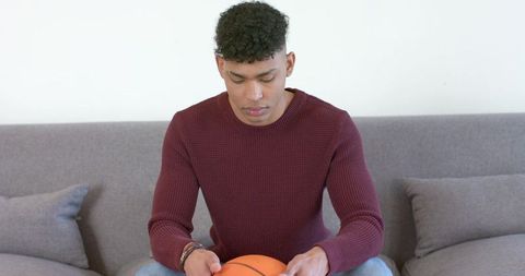 Contemplative young man sitting on sofa holding basketball in minimalist living room