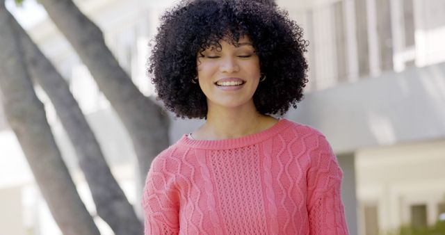 Young woman with curly hair wearing a cozy pink sweater and smiling in an outdoor setting. Ideal for lifestyle, fashion, and positive emotion concepts. Perfect for use in ads, blogs, and social media posts related to fashion, casual lifestyle, and happy moments.