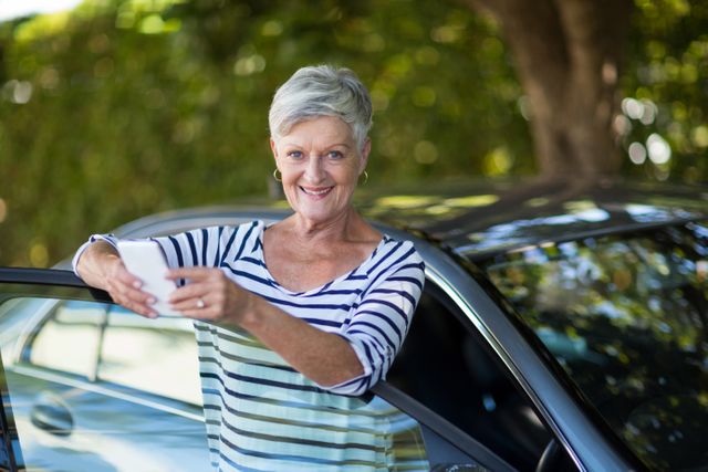 Senior woman holding phone while leaning on car door - Download Free Stock Photos Pikwizard.com