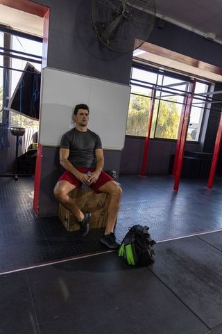 Tired athlete resting on plyo box after workout holding water bottle in industrial gym