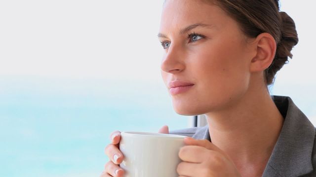 A businesswoman is drinking coffee while contemplatively gazing out at the sea. Her calm and relaxed posture suggests a peaceful moment, making it suitable for themes around mindfulness, relaxation, or balancing work and leisure. It can be used for business lifestyle promotions, work-from-home concepts, or emphasizing the benefits of taking breaks for mental clarity.