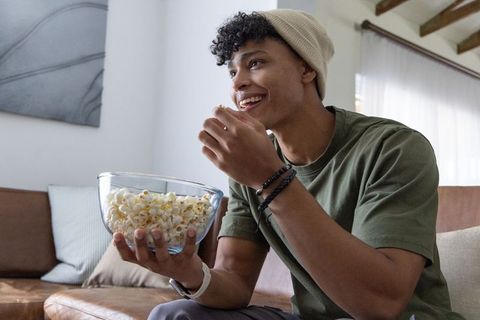 African american man wearing beanie watching movie and eating popcorn on sofa