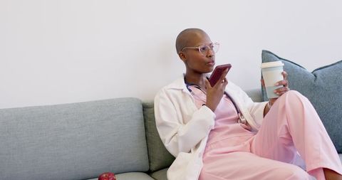 Clinician relaxing on couch in scrubs holding coffee and checking phone during break