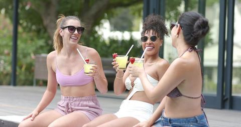 Three women laughing and toasting tropical cocktails on pool deck in summer swimwear