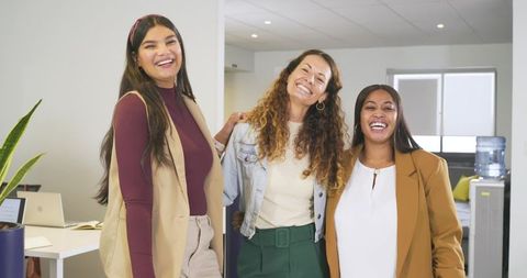 Three multicultural female coworkers smiling and posing in bright modern office workspace