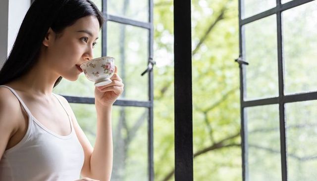Asian woman sipping tea from floral china cup beside black-paned metal window, serene