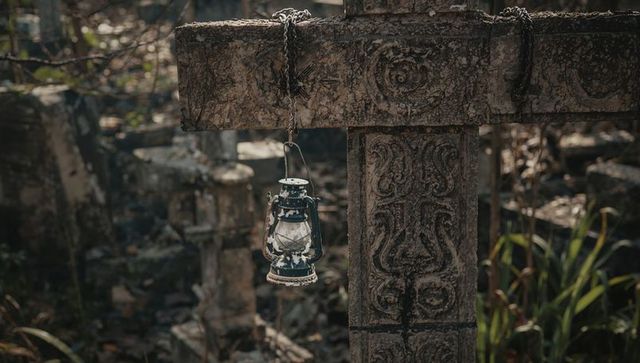Weathered lantern hanging from carved stone cross in overgrown graveyard ruin, moody forest