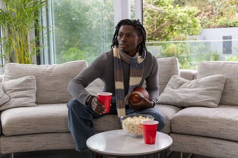 African american man watching football on modern sofa holding football and red cup