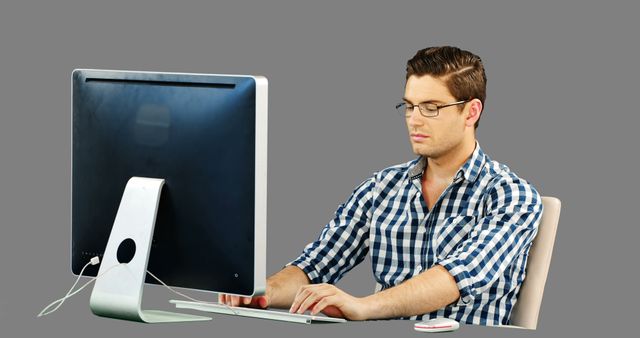 Young, male professional wearing glasses and plaid shirt is using desktop computer at work. Ideal for illustrating productivity, working from home, office technology, and professional focus.