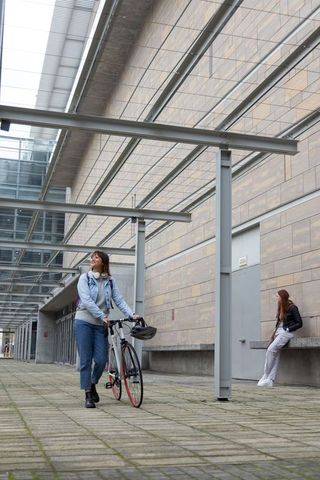 Young woman pushing bicycle through modern covered urban atrium with friend leaning on wall