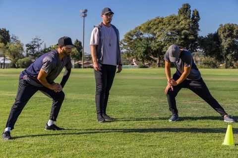 Coach leading male athletes through dynamic lateral stretches on grass field