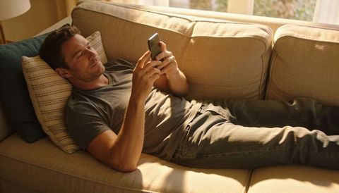 Casual man reclining on beige sofa using smartphone in warm sunlit living room