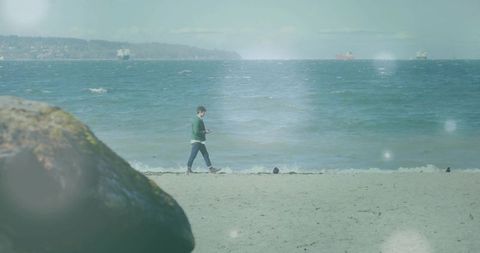 Man walking along shoreline checking smartphone with cargo ships on hazy ocean horizon