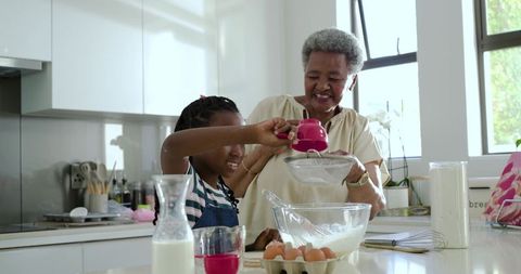 Grandmother guiding granddaughter sifting flour while baking in bright home kitchen