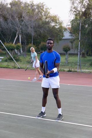 Male tennis player holding racket while partner practicing on outdoor hard court
