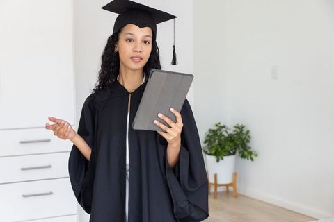 Young graduate presenting with tablet in bright home office wearing cap and gown