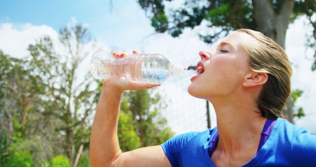 A young Caucasian woman is drinking water from a bottle outdoors, with copy space. Staying hydrated is essential during physical activities, especially in warm weather.