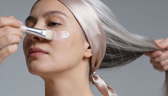 Midlife asian woman applying skincare cream with flat brush, satin headscarf, minimalist beauty