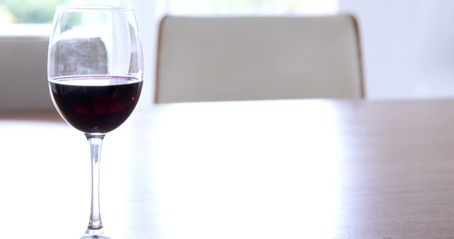 Glass of red wine placed on modern wooden table in a well-lit room with a minimalist design. Perfect for use in articles about wine tasting, interior design, elegance, and contemporary lifestyles.