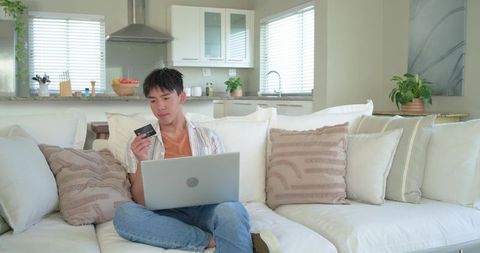 Asian man sitting on sofa using laptop holding credit card for online shopping at home