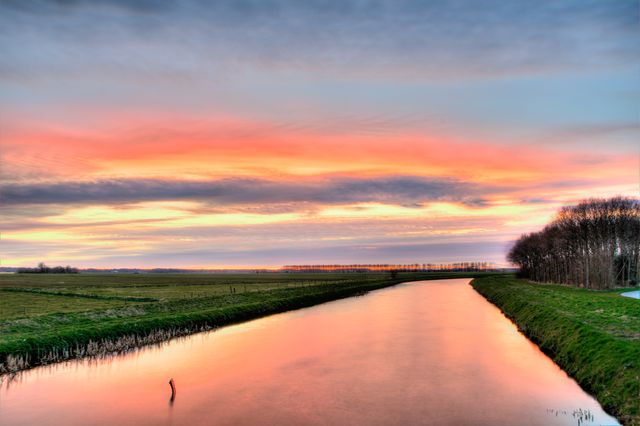 This image reveals a serene river reflecting vibrant colors of the sky during sunset. The scene captures the essence of tranquility with the calm water and expansive horizons. It can be used in travel brochures, relaxation and wellness promotions, landscape and nature-themed websites, or as a background image to evoke peace and beauty in design projects.