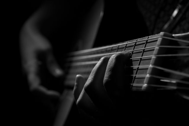 Close-up of guitarist's hand strumming acoustic guitar in low light creates dramatic and intimate ambiance. Black and white tones enhance focus on musician's technique and strings. Perfect for music-related content, posters, album covers, promotional materials, and articles on musicianship, practice, and performances.