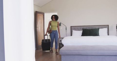 African american woman entering modern hotel bedroom pulling suitcase and smiling in casual outfit