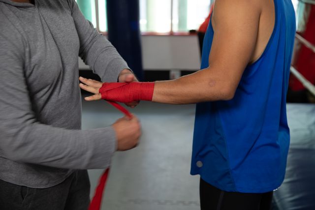This image shows a young Caucasian male boxer having his hands wrapped by a middle-aged Caucasian male trainer in a boxing ring. Ideal for use in articles or advertisements related to boxing, sports training, fitness, teamwork, and athletic preparation. It can also be used in promotional materials for gyms, boxing clubs, and sports coaching services.