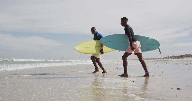 Two men walking along sandy beach carrying surfboards. Ideal for themes related to surfing, beach vacations, outdoor activities, and friendship. Useful for promotional materials for coastal destinations, surf schools, and recreational sports advertisements.