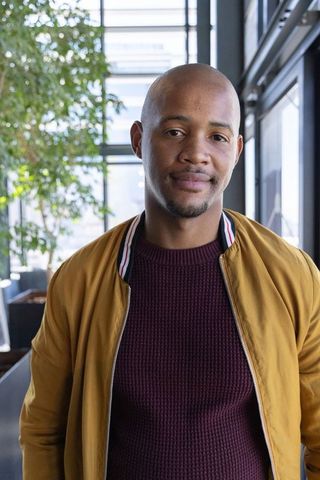 Confident african american man standing in modern atrium wearing mustard bomber jacket