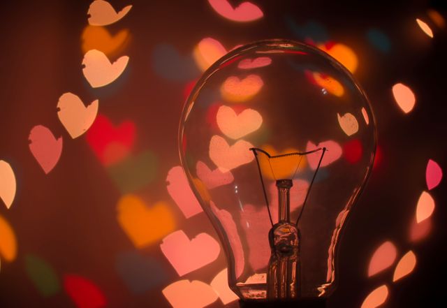 Colorful heart-shaped bokeh lights surround clear light bulb, creating romantic, dreamy atmosphere. Perfect for Valentine's Day, love-themed designs, romantic greeting cards, and wedding invitations. Can also be used for creative artwork, DIY decoration inspiration, and celebrating love.