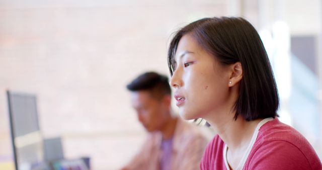 Asian woman in casual attire deeply focused on work, embodying dedication and engagement in a modern office environment. Man in the background appearing blurred adds a sense of depth and realism, highlighting modern workplace dynamics. Useful for depicting professionalism, dedication in tech-driven task settings, or teamwork in diverse work environments.