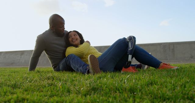 Diverse couple enjoying a joyful moment outside while sitting on grass. Woman with prosthetic leg smiling warmly. Suitable for themes related to love, inclusivity, companionship, overcoming challenges, happiness, and diversity.