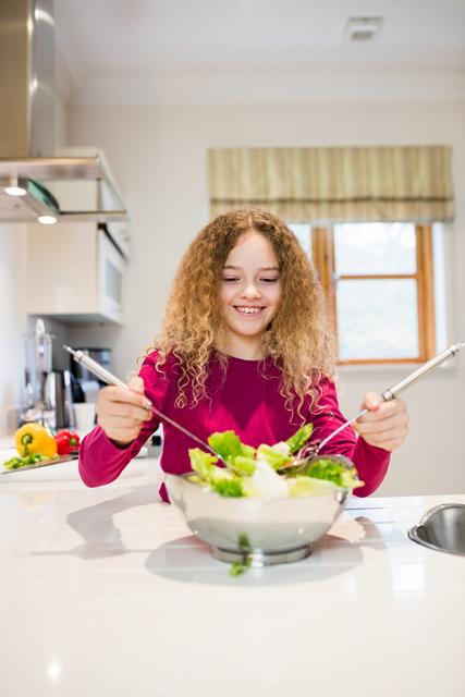Young girl with curly hair preparing a fresh salad in a modern kitchen. She is smiling and enjoying the process of cooking. This image can be used for promoting healthy eating habits, family activities, and cooking tutorials. Ideal for websites, blogs, and advertisements related to nutrition, family life, and home cooking.