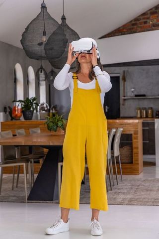 Middle-aged woman exploring virtual reality in bright open-plan kitchen wearing yellow overalls