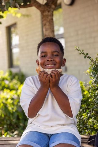 Smiling african american boy eating sandwich on bench sunny backyard white tee denim shorts