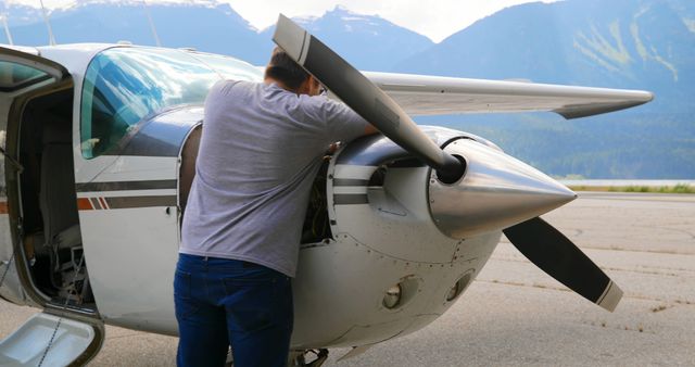 Mechanic examining propeller engine on small aircraft parked at airport outdoors. Ideal for use in content related to aviation, aircraft maintenance, engineering, transportation, pilot training, and safety inspection guides.