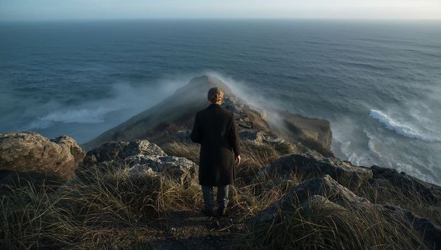 Solitary figure standing on windswept coastal cliff path overlooking misty ocean headland