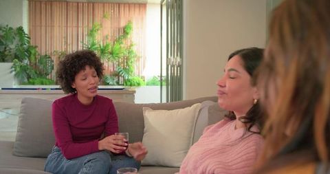 Diverse women chatting and sipping drinks on cozy gray sofa with indoor plants